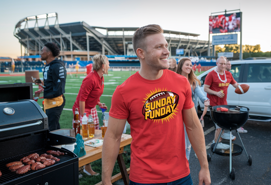 A man in a red t-shirt with Sunday Funday design on the front of the t-shirt. There are people in the background with footballs, and grilling food.